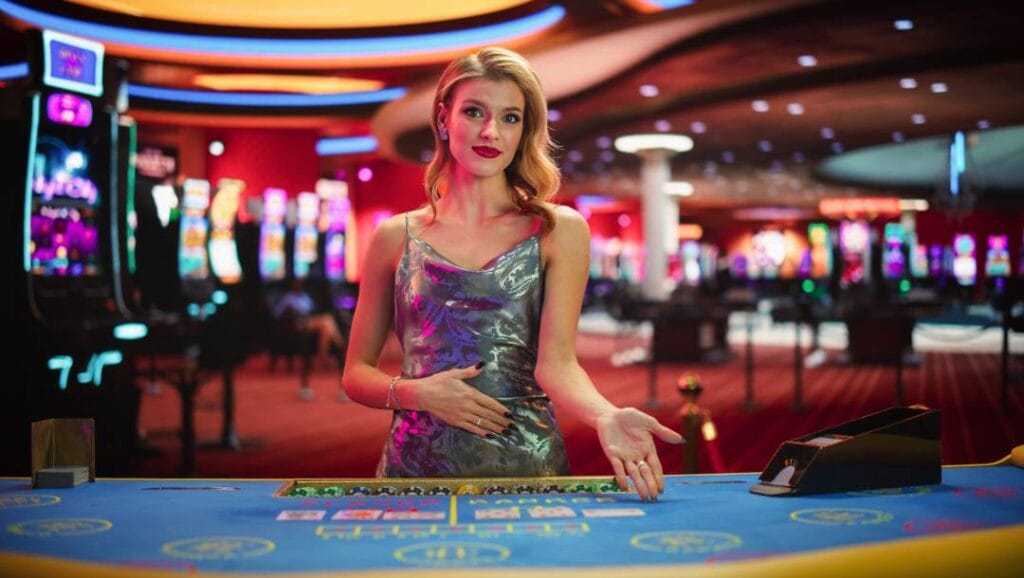 A blonde casino dealer in a silver dress gesturing to a player to have a seat at a baccarat table in a casino. Brightly lit slot game machines are out of focus behind her.