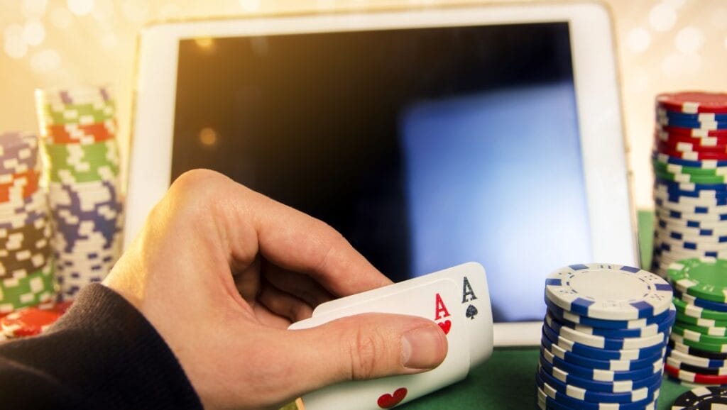 A hand holding ace poker cards in front of a tablet device and casino chips on a green table.