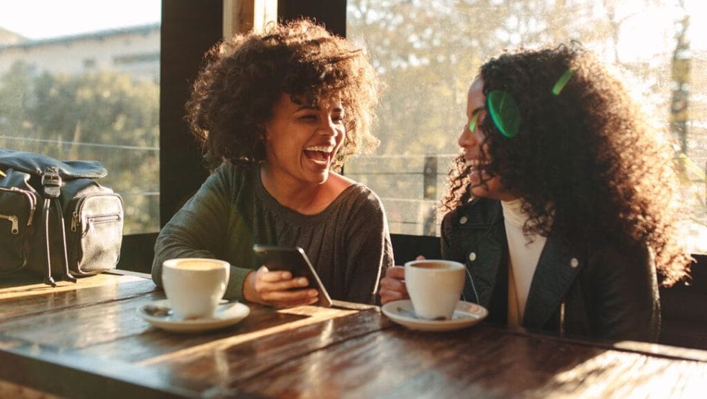 Two female friends sitting at a table at a coffee shop, looking at a smartphone screen and laughing.