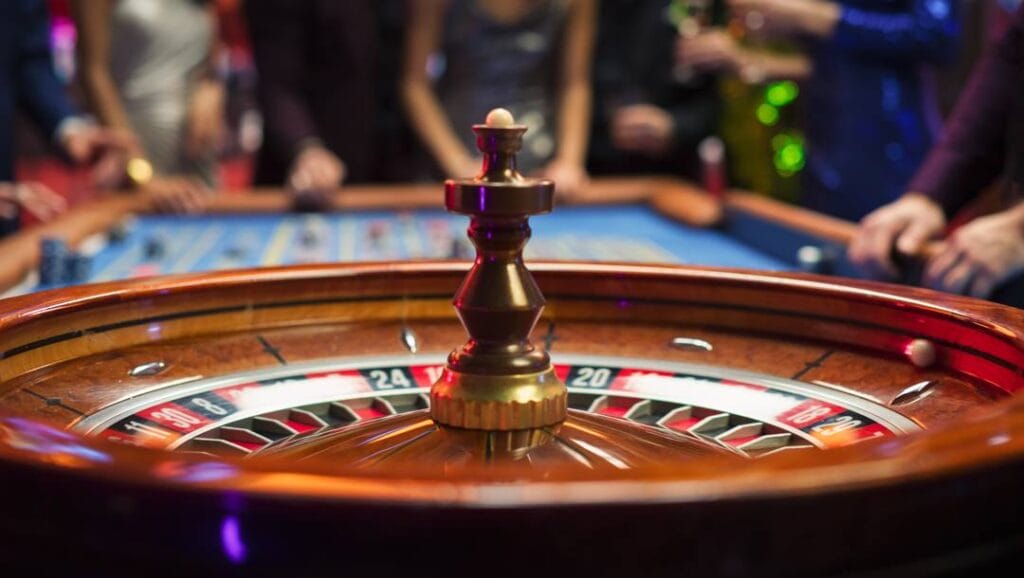 A close-up of a roulette wheel with a roulette ball spinning inside it. The rest of the roulette table is visible in the background. There are many gamblers waiting around it.
